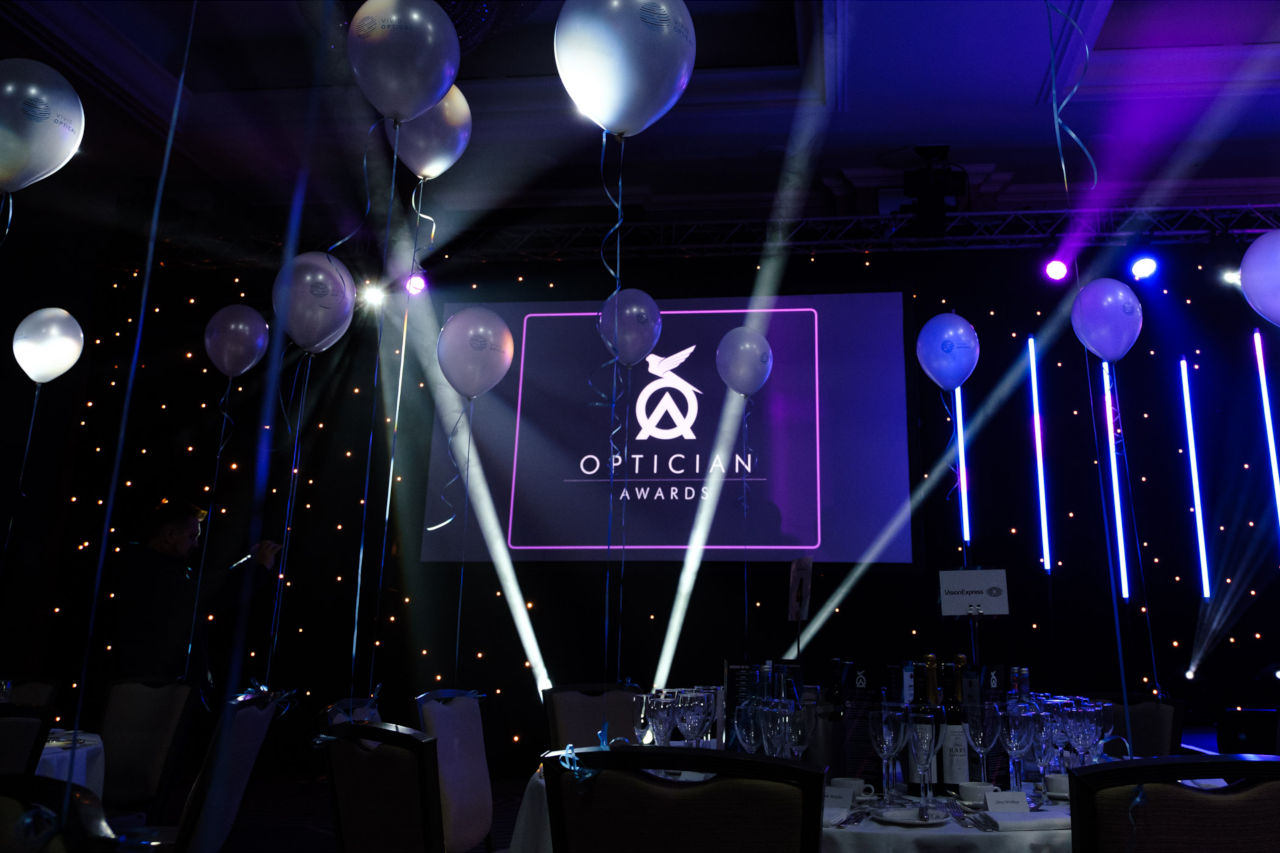 Balloons and champagne glasses on tables in a darkened room, showing the Optician Awards logo on a screen